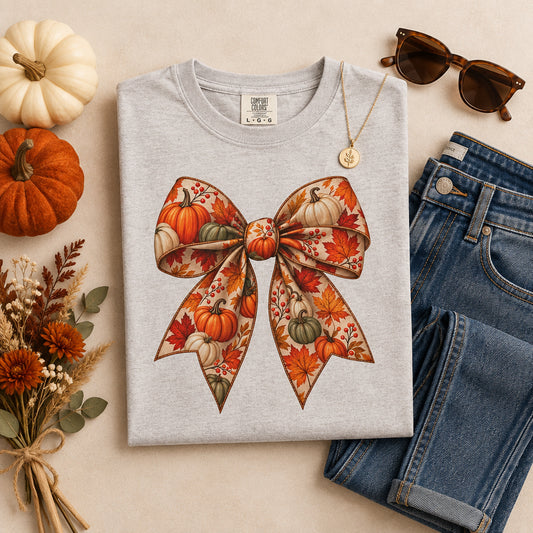 A white short-sleeve crewneck t-shirt featuring a large graphic of a bow patterned with orange and green pumpkins and autumn leaves.