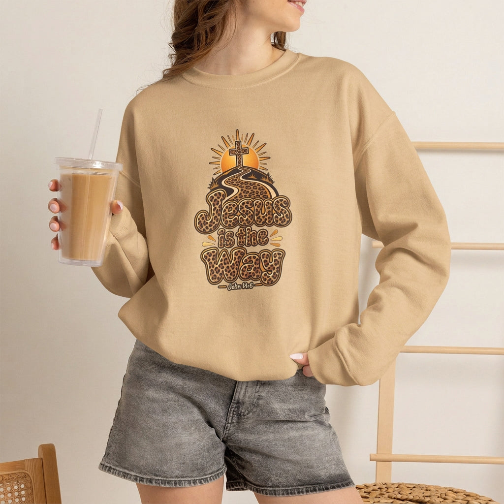 A woman wearing a white crewneck sweatshirt with a graphic print that reads 'Jesus is the Way' in leopard-print lettering with a sun and tree design. She is holding an iced coffee and wearing gray denim shorts in a brightly lit indoor setting.