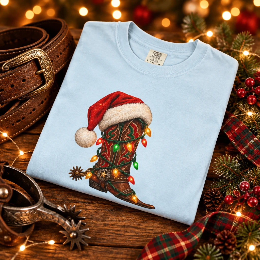 A light blue folded crewneck t-shirt featuring a graphic of a brown cowboy boot decorated with a red Santa hat and colorful Christmas lights.
