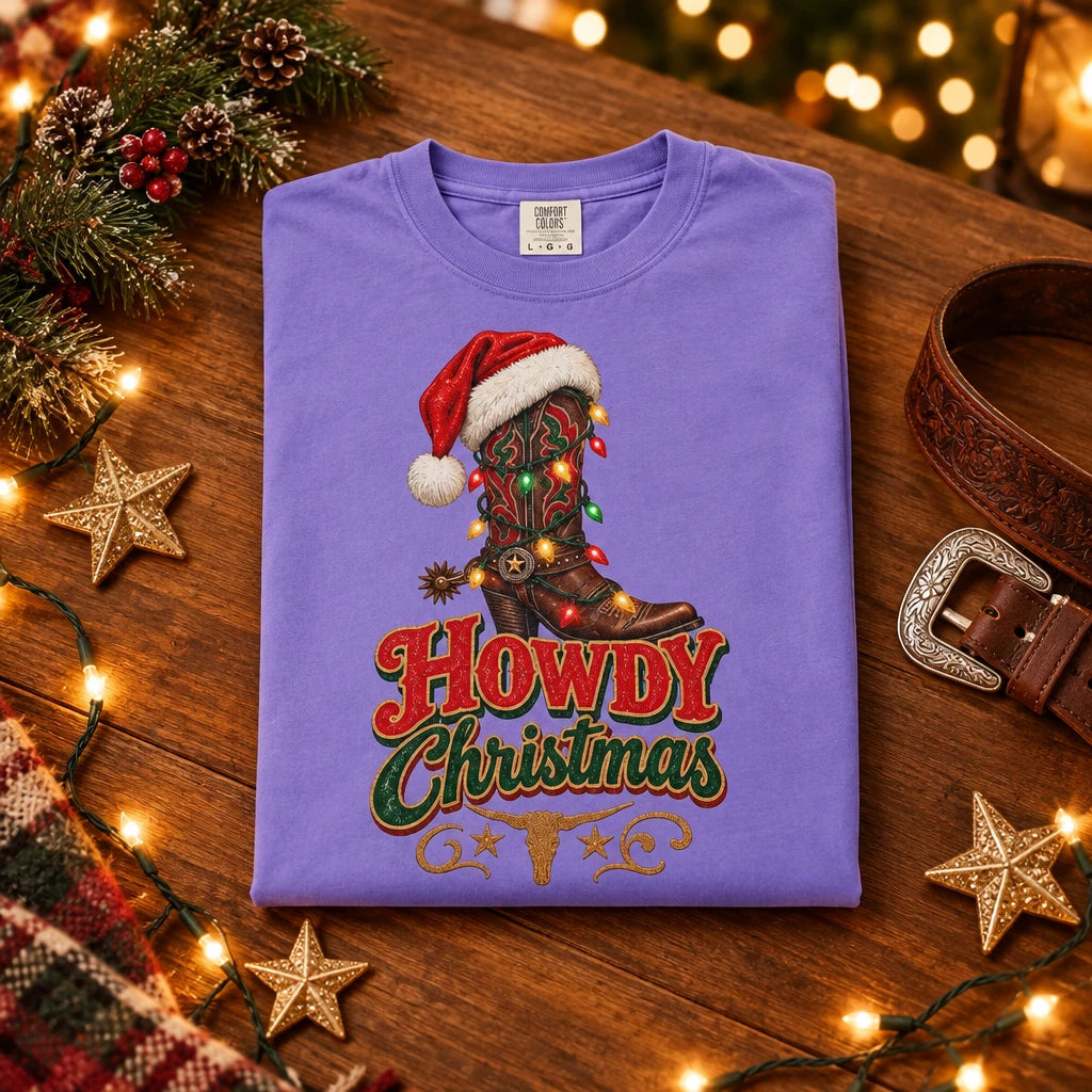 A purple short-sleeve crewneck t-shirt featuring a graphic of a cowboy boot decorated with Christmas lights and a Santa hat, above the text 'Howdy Christmas' in red and green.