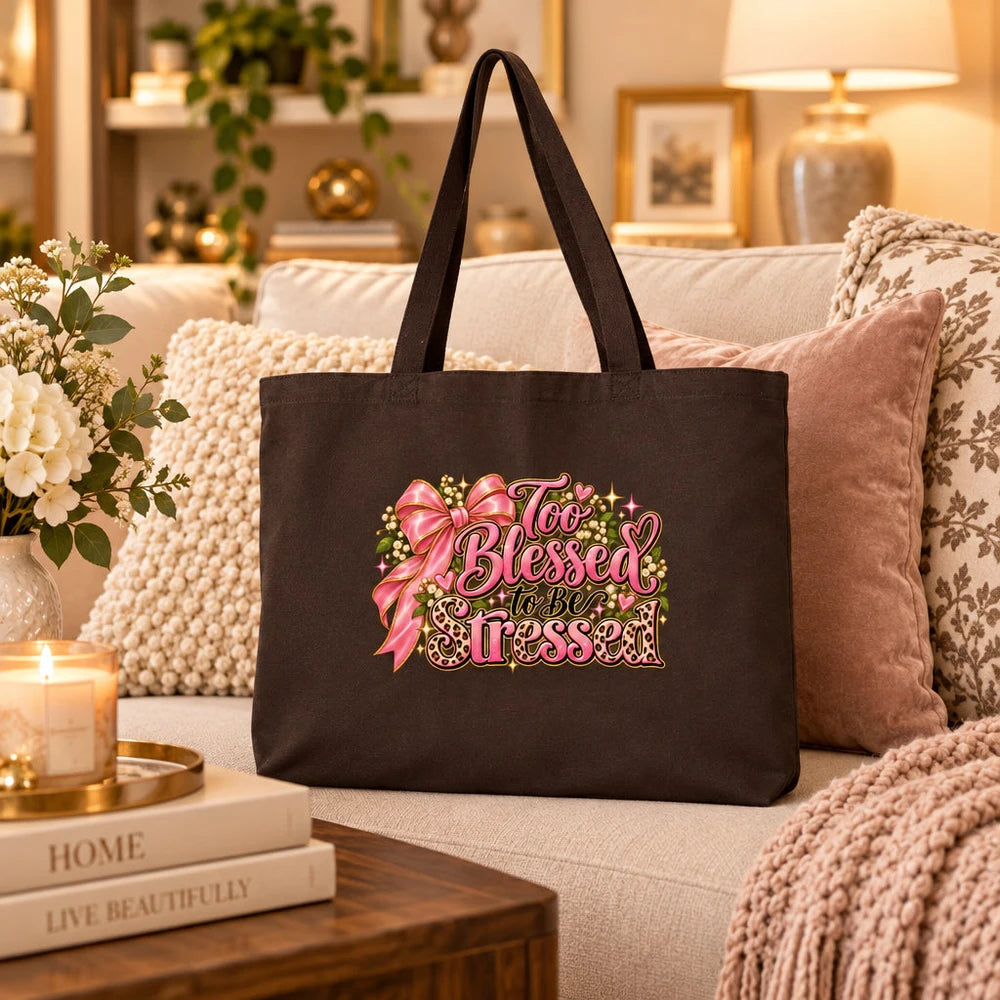 A black canvas tote bag with a pink and green 'Too Blessed to Be Poor' graphic, styled on a cozy beige sofa with decorative pillows and candles in a warm living room setting.