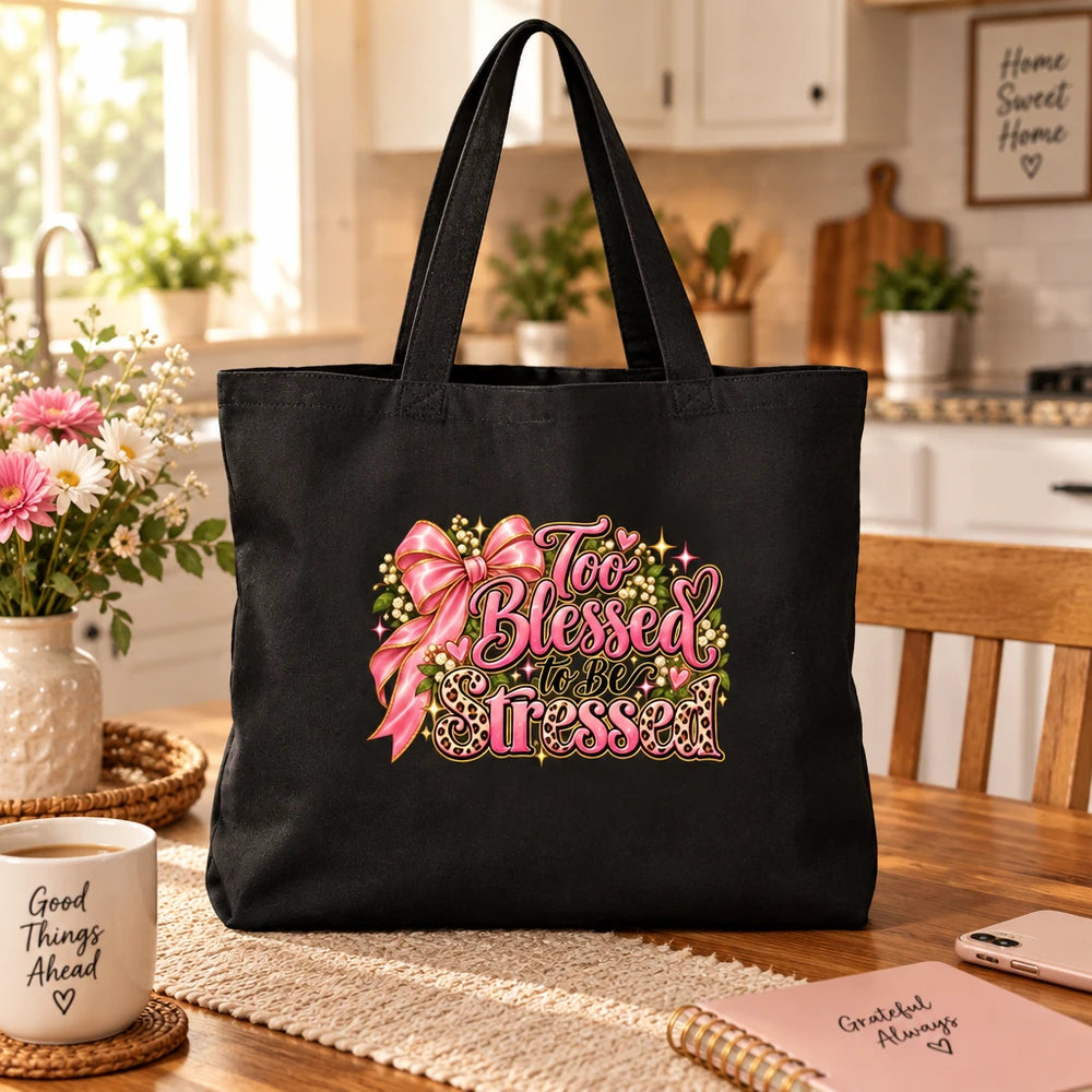 A black canvas tote bag with a pink and gold graphic reading 'Too Blessed to Be Less' sits on a wooden dining table in a warm, sunlit kitchen setting.
