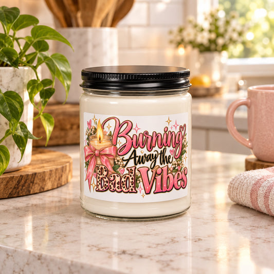 A glass jar candle with a black metal lid and a white label that reads 'Burning Away the Bad Vibes' in pink and black script, decorated with a pink bow, candle flame, and floral elements.