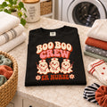 A black short-sleeve crewneck t-shirt featuring a retro-style graphic on the front with the text 'BOO BOO CREW ER NURSE' and three cute cartoon ghosts dressed as nurses holding stethoscopes, shown against a plain white background.